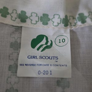May include: Close-up of a white fabric with a green clover pattern and a tag. The tag features the Girl Scouts logo, the number 10, and the text "GIRL SCOUTS" and "SEE REVERSE FOR CARE & CONTENTS".