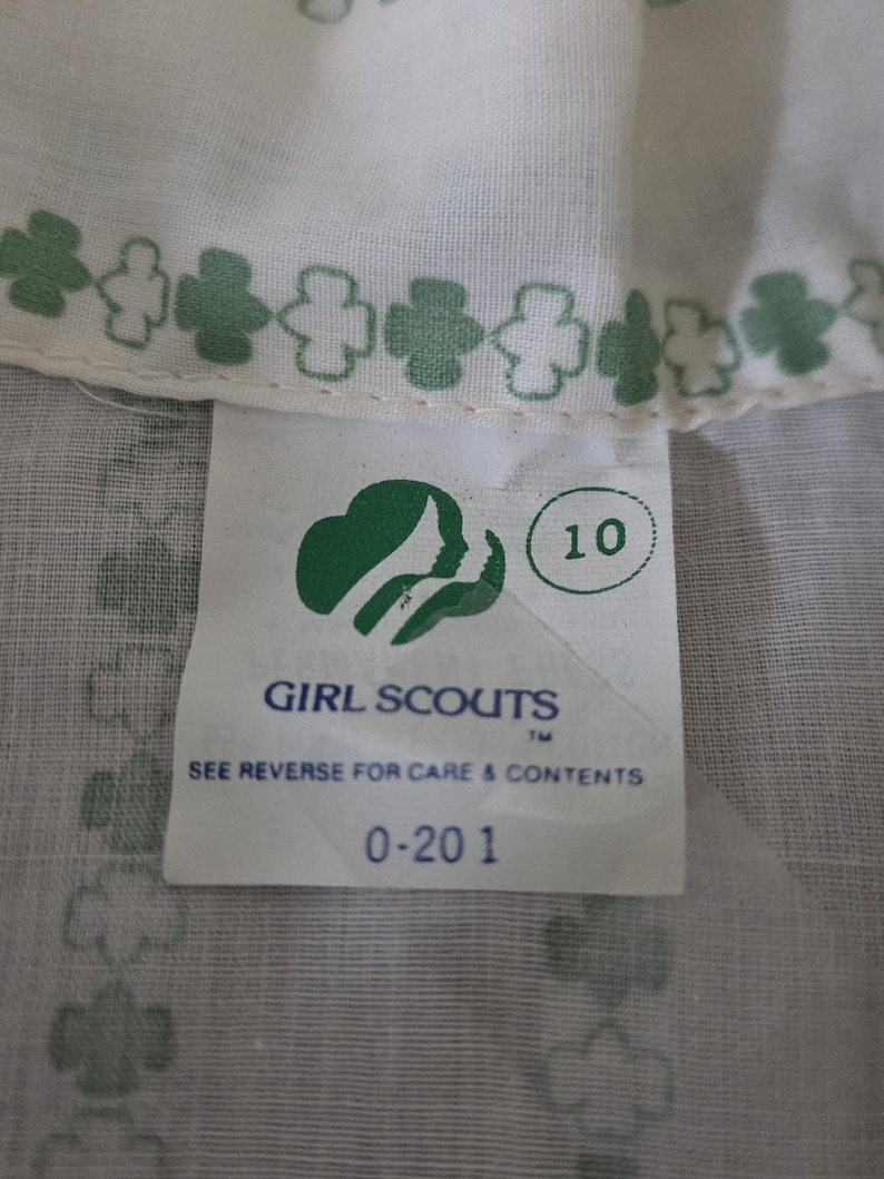 May include: Close-up of a white fabric with a green clover pattern and a tag. The tag features the Girl Scouts logo, the number 10, and the text "GIRL SCOUTS" and "SEE REVERSE FOR CARE & CONTENTS".