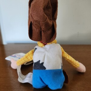 Op de afbeelding: Een pluche speelgoed van Woody uit de Toy Story-franchise. Woody draagt zijn kenmerkende bruine cowboyhoed, een geel en bruin geruit overhemd, een wit vest met een zwart-wit koe patroon, en een blauwe spijkerbroek.