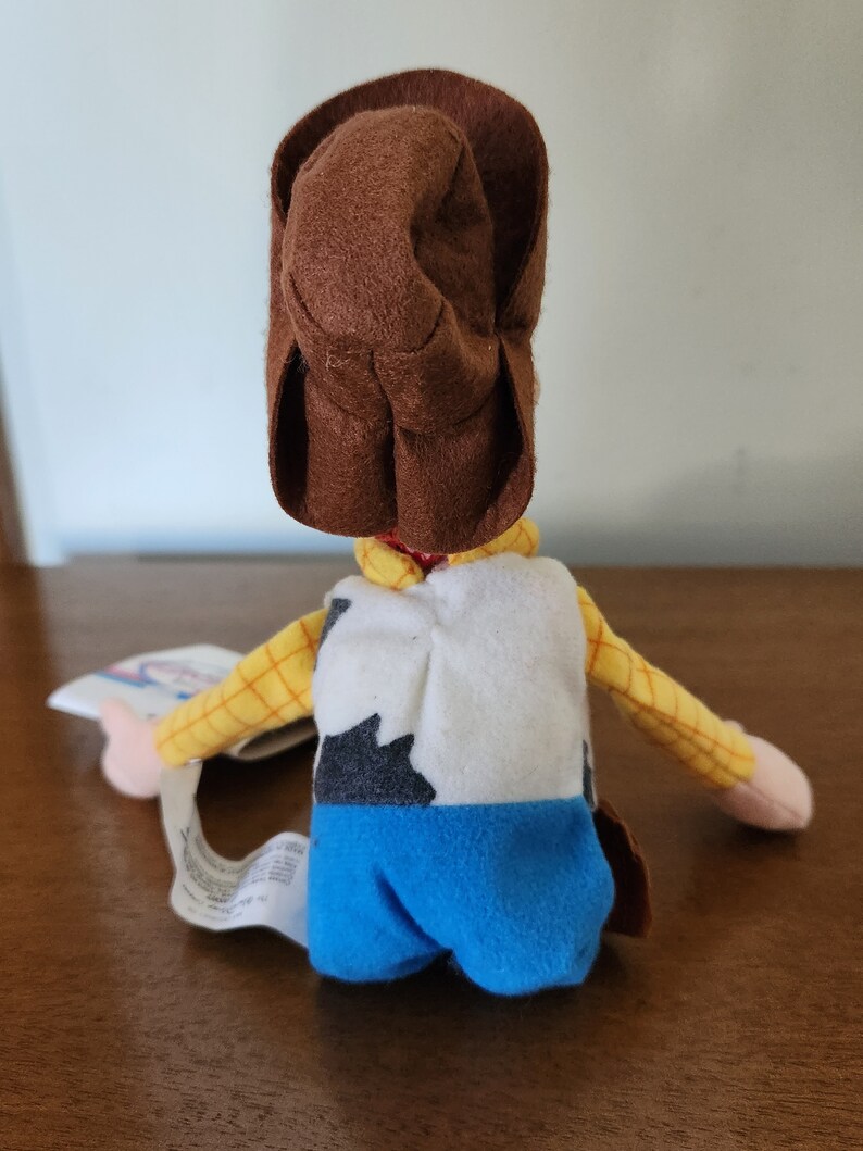 Op de afbeelding: Een pluche speelgoed van Woody uit de Toy Story-franchise. Woody draagt zijn kenmerkende bruine cowboyhoed, een geel en bruin geruit overhemd, een wit vest met een zwart-wit koe patroon, en een blauwe spijkerbroek.
