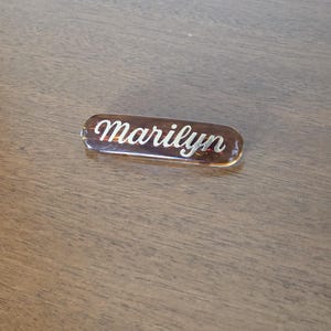 May include: A rectangular name tag with the name "Marilyn" in white script. The tag has a brown, amber-like appearance and is set against a wooden surface.