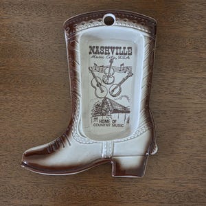 May include: A ceramic ashtray shaped like a cowboy boot, featuring a brown and cream color scheme. The ashtray has the text "Nashville Music City, U.S.A." and illustrations of musical instruments. The words "Home of Country Music" are also visible.