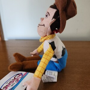Op de afbeelding: Een pluche speelgoed van Woody uit de Toy Story-franchise. Woody draagt zijn kenmerkende bruine hoed, geel en blauw shirt en bruine laarzen. Hij zit met gekruiste armen en kijkt naar rechts. Het speelgoed is voorzien van een "Disney Store"-label.