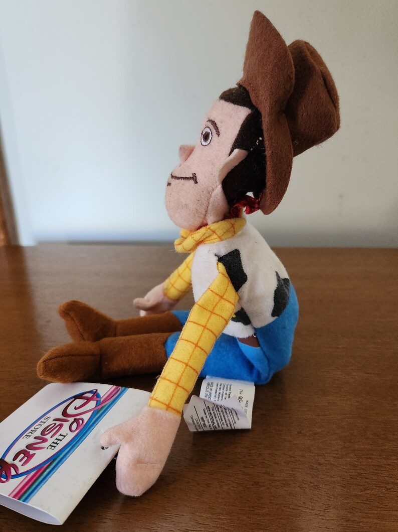 Op de afbeelding: Een pluche speelgoed van Woody uit de Toy Story-franchise. Woody draagt zijn kenmerkende bruine hoed, geel en blauw shirt en bruine laarzen. Hij zit met gekruiste armen en kijkt naar rechts. Het speelgoed is voorzien van een "Disney Store"-label.