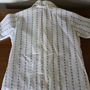 May include: White button-up shirt with vertical stripes of green clover-like shapes. The shirt has short sleeves and a collar. The pattern is repeated on the collar and sleeve edges. The shirt is made of a lightweight fabric.