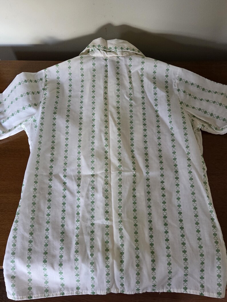 May include: White button-up shirt with vertical stripes of green clover-like shapes. The shirt has short sleeves and a collar. The pattern is repeated on the collar and sleeve edges. The shirt is made of a lightweight fabric.