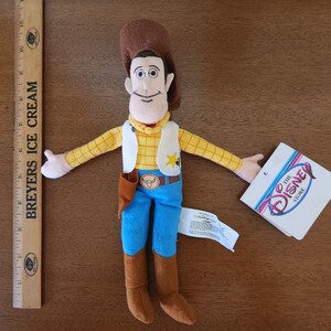 Op de afbeelding: Een pluche speelgoed van Woody uit de Toy Story-franchise. Woody draagt zijn kenmerkende bruine hoed, gele vest en blauwe jeans. Hij heeft een bruine riem met een sheriffster en een speelgoed stierenkop gesp. Het speelgoed houdt zijn armen uitgestrekt. Het speelgoed zit op een houten oppervlak. Het label op het speelgoed luidt "The Disney Store."