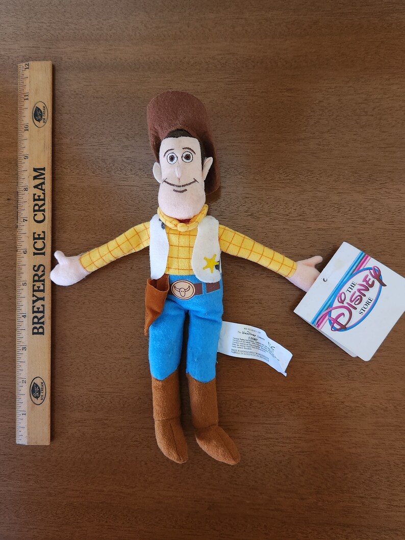 Op de afbeelding: Een pluche speelgoed van Woody uit de Toy Story-franchise. Woody draagt zijn kenmerkende bruine hoed, gele vest en blauwe jeans. Hij heeft een bruine riem met een sheriffster en een speelgoed stierenkop gesp. Het speelgoed houdt zijn armen uitgestrekt. Het speelgoed zit op een houten oppervlak. Het label op het speelgoed luidt "The Disney Store."