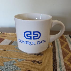 May include: White ceramic mug with a blue logo that reads "CONTROL DATA". The mug has a curved handle and a slightly flared rim. The mug is sitting on a patterned, tan-colored surface.