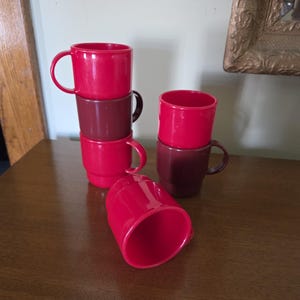 May include: A collection of red and maroon plastic mugs stacked and arranged on a wooden surface. The mugs have a simple, cylindrical design with curved handles. One mug is lying on its side. The background includes a wall and a framed object.