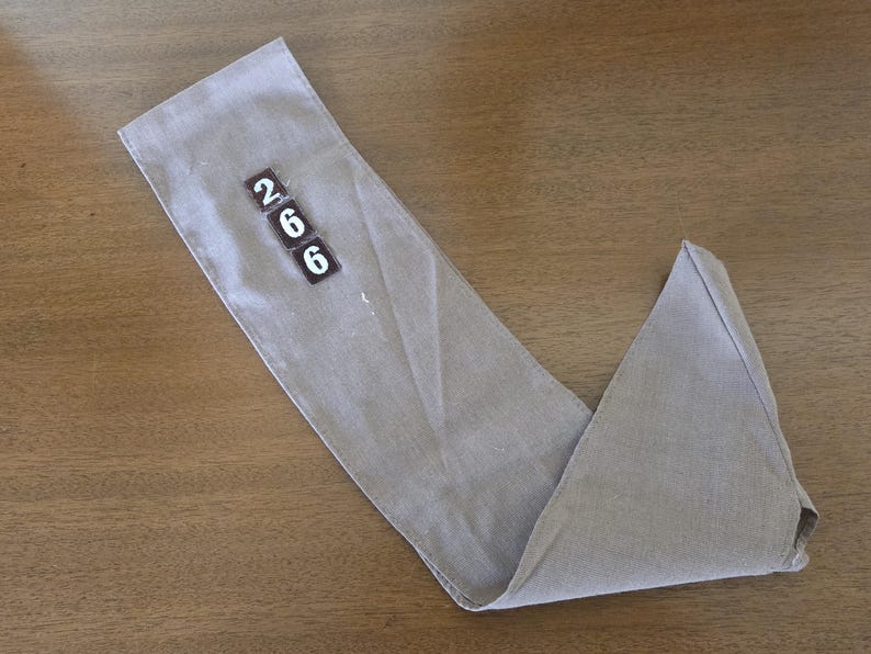 May include: A folded, light gray fabric item with the numbers "266" printed on a small rectangular patch. The fabric is lying on a wooden surface, with the item folded into a V-shape.