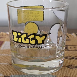 May include: Clear glass tumbler featuring the word "Ziggy" in yellow and black lettering. The tumbler has cartoon illustrations and text bubbles. The artist's signature, "Tom Wilson," is visible below the logo.