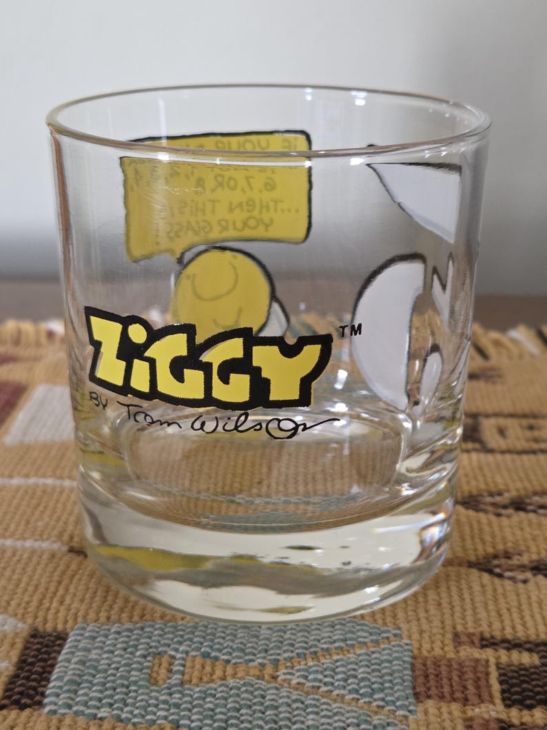 May include: Clear glass tumbler featuring the word "Ziggy" in yellow and black lettering. The tumbler has cartoon illustrations and text bubbles. The artist's signature, "Tom Wilson," is visible below the logo.