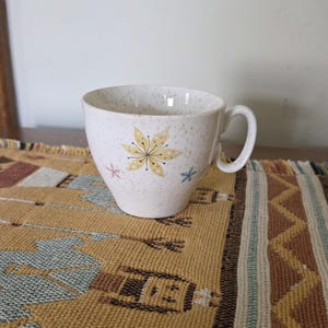 May include: A speckled white ceramic teacup with a handle, decorated with a yellow floral design and small pink and blue accents. The cup sits on a woven, patterned table runner.