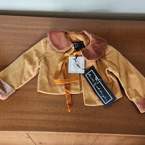 May include: A mustard yellow corduroy jacket with a brown velvet collar and matching brown velvet trim on the cuffs. The jacket has a ribbon tied at the front and a small tag that says "Petite & Petite".