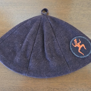 May include: A dark purple, triangular hat with a small loop at the top. The hat features a circular patch with an orange figure and a blue border. The hat is made of a soft, felt-like material.