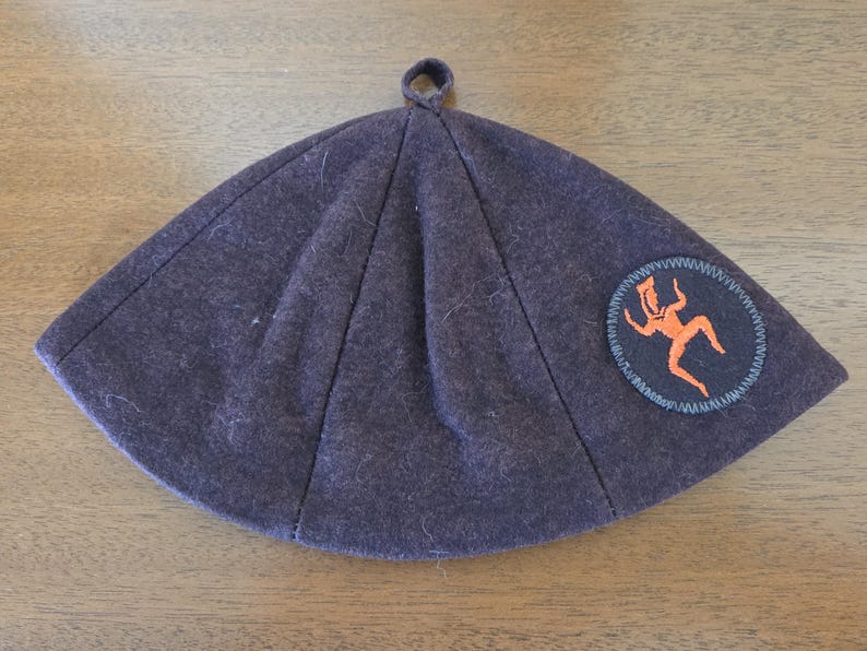 May include: A dark purple, triangular hat with a small loop at the top. The hat features a circular patch with an orange figure and a blue border. The hat is made of a soft, felt-like material.