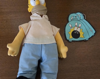 May include: A Homer Simpson plush doll with a yellow head, tan shirt, blue pants, and black shoes. A bowling bag tag is next to the doll. The tag is light blue with a bowling ball and pins.