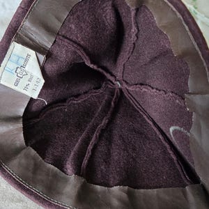 May include: Inside view of a vintage Girl Scout hat. The hat is a deep burgundy color with a brown leather band around the edge. A tag inside reads "Girl Scouts" and "70% Wool".