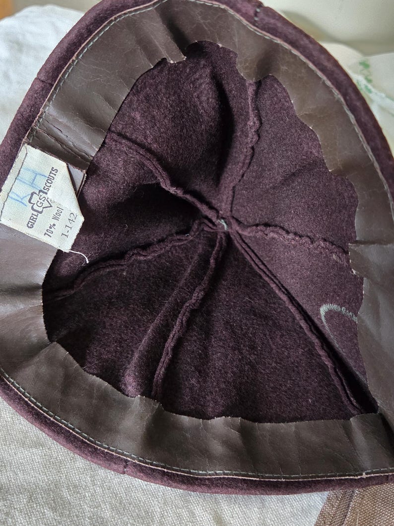 May include: Inside view of a vintage Girl Scout hat. The hat is a deep burgundy color with a brown leather band around the edge. A tag inside reads "Girl Scouts" and "70% Wool".