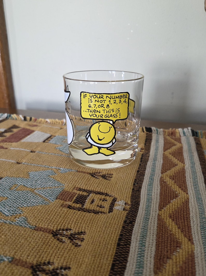 May include: A clear glass with a cartoon character and the text "IF YOUR NUMBER IS NOT 1,2,3,4, 6,7, OR 8...THEN THIS IS YOUR GLASS!". The glass is cylindrical and sits on a patterned surface.