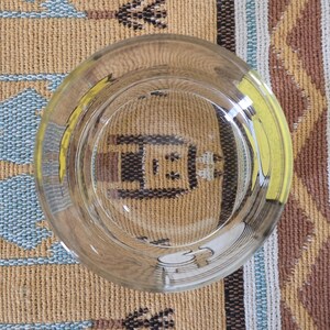 May include: Clear glass ashtray with a yellow accent, viewed from above. The ashtray reflects the pattern of the surface it rests on, a woven textile with tan, blue, and brown colors. The ashtray is circular and has a wide rim.