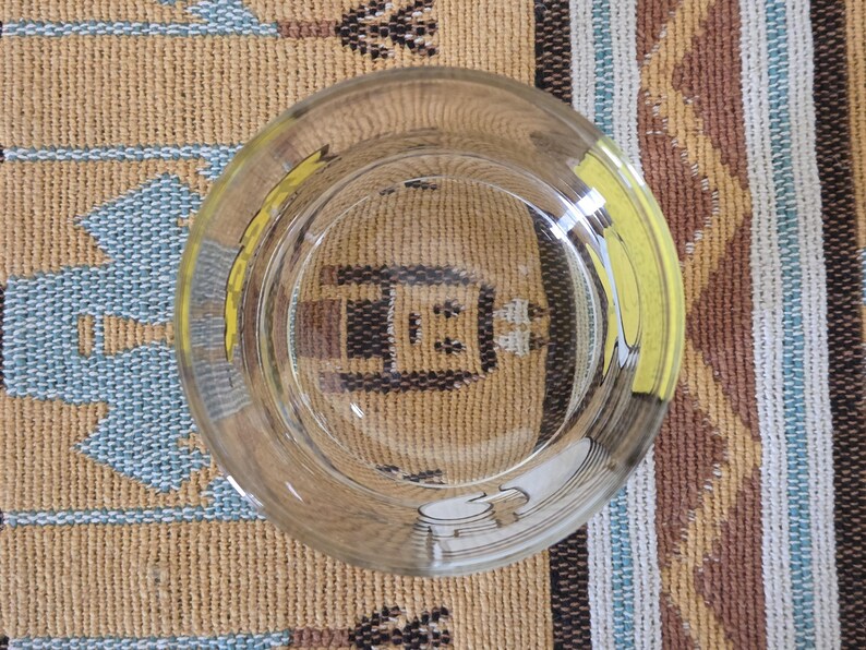May include: Clear glass ashtray with a yellow accent, viewed from above. The ashtray reflects the pattern of the surface it rests on, a woven textile with tan, blue, and brown colors. The ashtray is circular and has a wide rim.