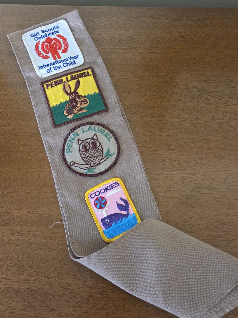 May include: A tan Girl Scout sash with four colorful patches. The patches include text and images of a red figure, a donkey, an owl, and a whale with the word "COOKIES".