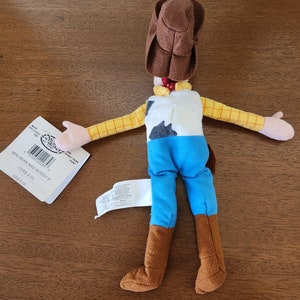 Op de afbeelding: Een bruin en geel pluche speelgoed van Woody uit de Toy Story-franchise. Het speelgoed draagt een bruine cowboyhoed, een blauw shirt en bruine laarzen. Het speelgoed ligt op zijn rug met zijn armen uitgestrekt. Het speelgoed heeft een label aan zijn arm dat "Mini Bean Bag Woody 8"  CONS 9.00 US$ 8.00" zegt.