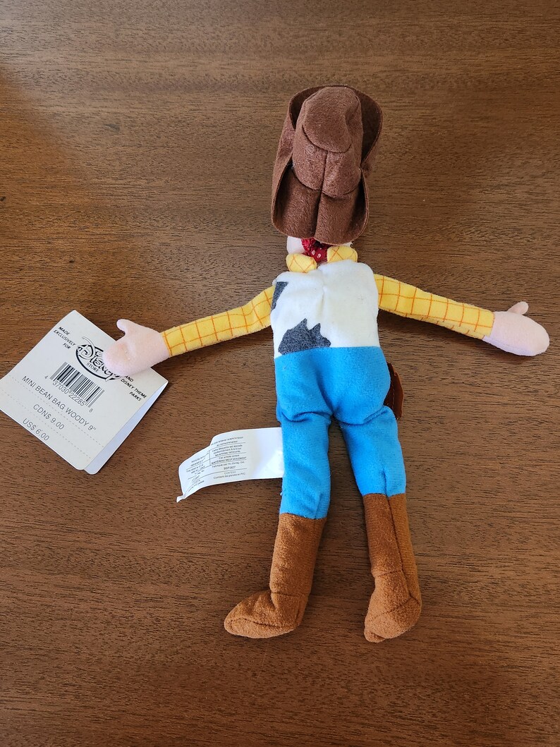 Op de afbeelding: Een bruin en geel pluche speelgoed van Woody uit de Toy Story-franchise. Het speelgoed draagt een bruine cowboyhoed, een blauw shirt en bruine laarzen. Het speelgoed ligt op zijn rug met zijn armen uitgestrekt. Het speelgoed heeft een label aan zijn arm dat "Mini Bean Bag Woody 8"  CONS 9.00 US$ 8.00" zegt.