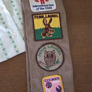 May include: A tan Girl Scout sash with several embroidered patches. Patches include "Girl Scouts Celebrate," "International Year of the Child," "Penn Laurel," and a patch with a rabbit and the number "79."