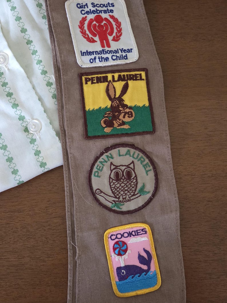 May include: A tan Girl Scout sash with several embroidered patches. Patches include "Girl Scouts Celebrate," "International Year of the Child," "Penn Laurel," and a patch with a rabbit and the number "79."