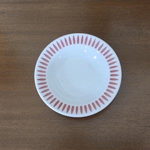 May include: White ceramic bowl with a pink, repeating, teardrop-shaped pattern around the rim. The bowl is centered on a wooden surface. The bowl is likely for serving food.