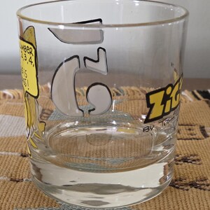 May include: Clear glass tumbler with a large number 6 in the center, outlined in black and filled with a light gray color. The word "Zit" is in yellow and black on the side. A yellow speech bubble with text is on the left.