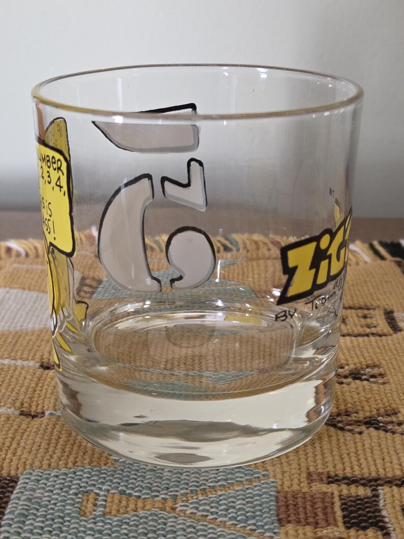 May include: Clear glass tumbler with a large number 6 in the center, outlined in black and filled with a light gray color. The word "Zit" is in yellow and black on the side. A yellow speech bubble with text is on the left.