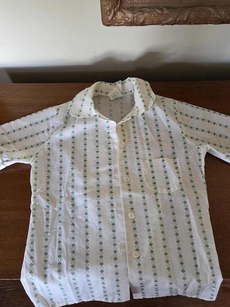 May include: White button-down shirt with a green clover pattern. The shirt has a collar, a chest pocket, and is buttoned up. The pattern is vertical stripes of green clovers.