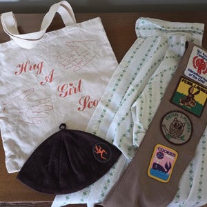 May include: An assortment of Girl Scout items, including a white tote bag with the text "Hug A Girl Scout", a brown hat, a uniform shirt, and a sash with patches. The sash has patches with images of a rabbit, an owl, and a whale.