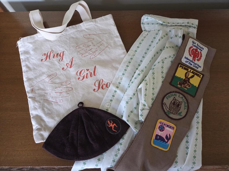 May include: An assortment of Girl Scout items, including a white tote bag with the text "Hug A Girl Scout", a brown hat, a uniform shirt, and a sash with patches. The sash has patches with images of a rabbit, an owl, and a whale.