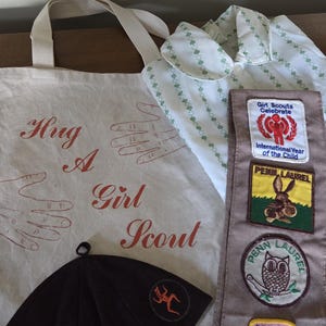 May include: An assortment of Girl Scout items, including a canvas tote bag with the text "Hug A Girl Scout", a brown hat, a white shirt with green clover patterns, and a sash with patches.