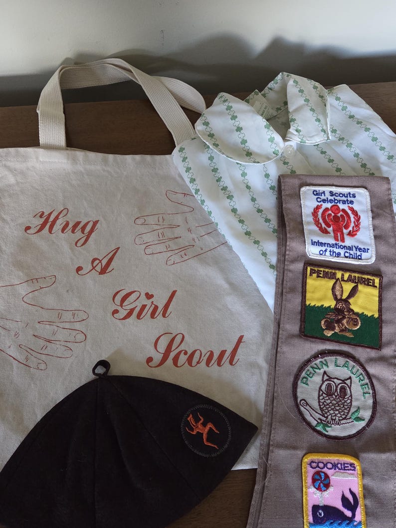 May include: An assortment of Girl Scout items, including a canvas tote bag with the text "Hug A Girl Scout", a brown hat, a white shirt with green clover patterns, and a sash with patches.