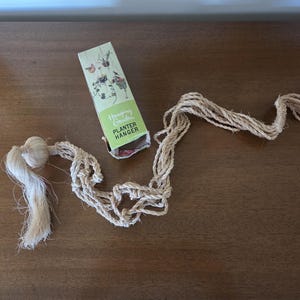 May include: A natural fiber macrame plant hanger with a decorative tassel and a box labeled "Hanging Garden Planter Hanger". The box is light green with illustrations of plants and birds.
