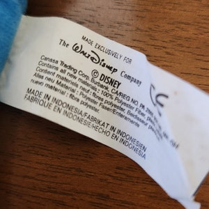 Op de afbeelding: Een wit label met zwarte tekst die "Exclusief gemaakt voor Walt Disney Company" en "&copy;DISNEY" aangeeft. Het label bevat ook informatie over de materialen die in het product zijn gebruikt, die 100% polyester zijn. Het label geeft aan dat het product in Indonesi&euml; is gemaakt.