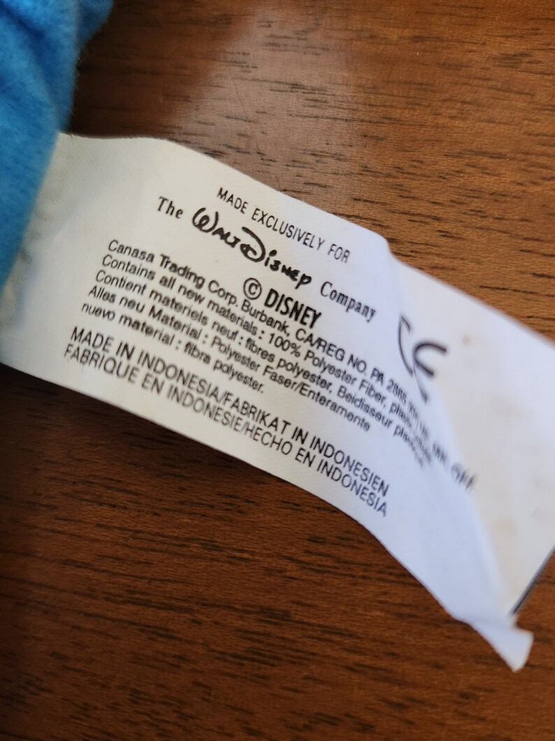 Op de afbeelding: Een wit label met zwarte tekst die "Exclusief gemaakt voor Walt Disney Company" en "&copy;DISNEY" aangeeft. Het label bevat ook informatie over de materialen die in het product zijn gebruikt, die 100% polyester zijn. Het label geeft aan dat het product in Indonesi&euml; is gemaakt.