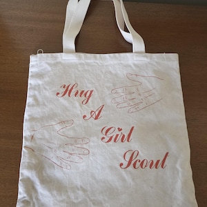 May include: Off-white canvas tote bag with cream-colored handles. The bag features the words "Hug A Girl Scout" in red script, along with red hand illustrations. The bag is made of a durable fabric.