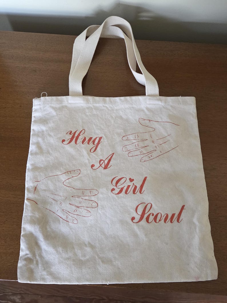 May include: Off-white canvas tote bag with cream-colored handles. The bag features the words "Hug A Girl Scout" in red script, along with red hand illustrations. The bag is made of a durable fabric.