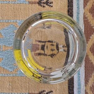 May include: A clear, round glass with a thick base, reflecting the patterned surface beneath. The glass has a slight greenish tint and is positioned over a tan, blue, and yellow patterned textile.