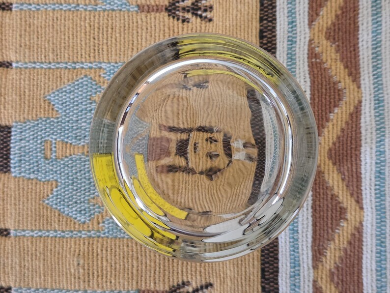 May include: A clear, round glass with a thick base, reflecting the patterned surface beneath. The glass has a slight greenish tint and is positioned over a tan, blue, and yellow patterned textile.
