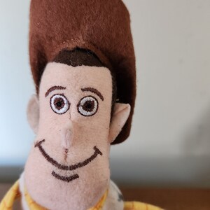 Op de afbeelding: Een pluche speelgoed van Woody uit de Toy Story-franchise. Het speelgoed draagt een bruine cowboyhoed, een gele vest en een blauw shirt. Het speelgoed heeft een bruine snor en een vriendelijke glimlach.