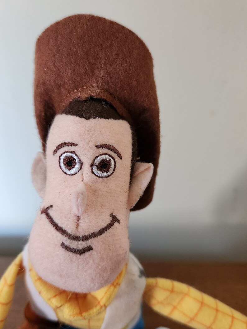 Op de afbeelding: Een pluche speelgoed van Woody uit de Toy Story-franchise. Het speelgoed draagt een bruine cowboyhoed, een gele vest en een blauw shirt. Het speelgoed heeft een bruine snor en een vriendelijke glimlach.