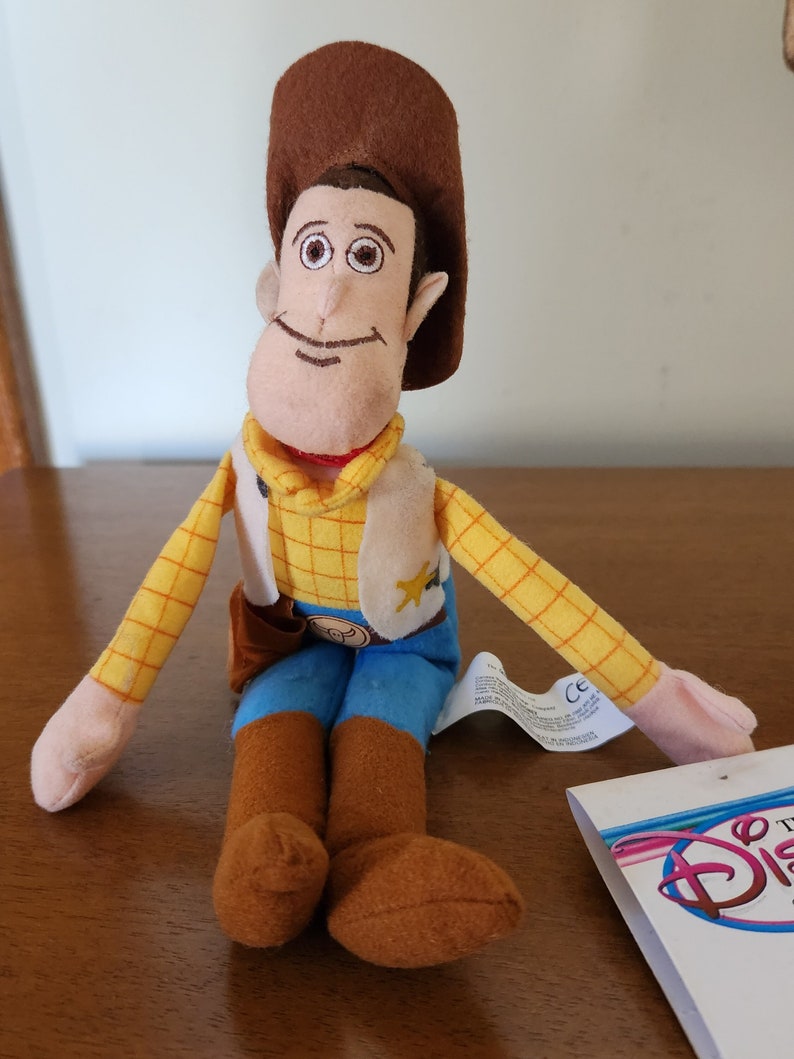 Op de afbeelding: Een pluche speelgoed van Woody uit de Toy Story-franchise. Hij draagt zijn kenmerkende cowboyhoed, vest en laarzen. Hij zit met gekruiste benen en heeft een vriendelijke glimlach op zijn gezicht.